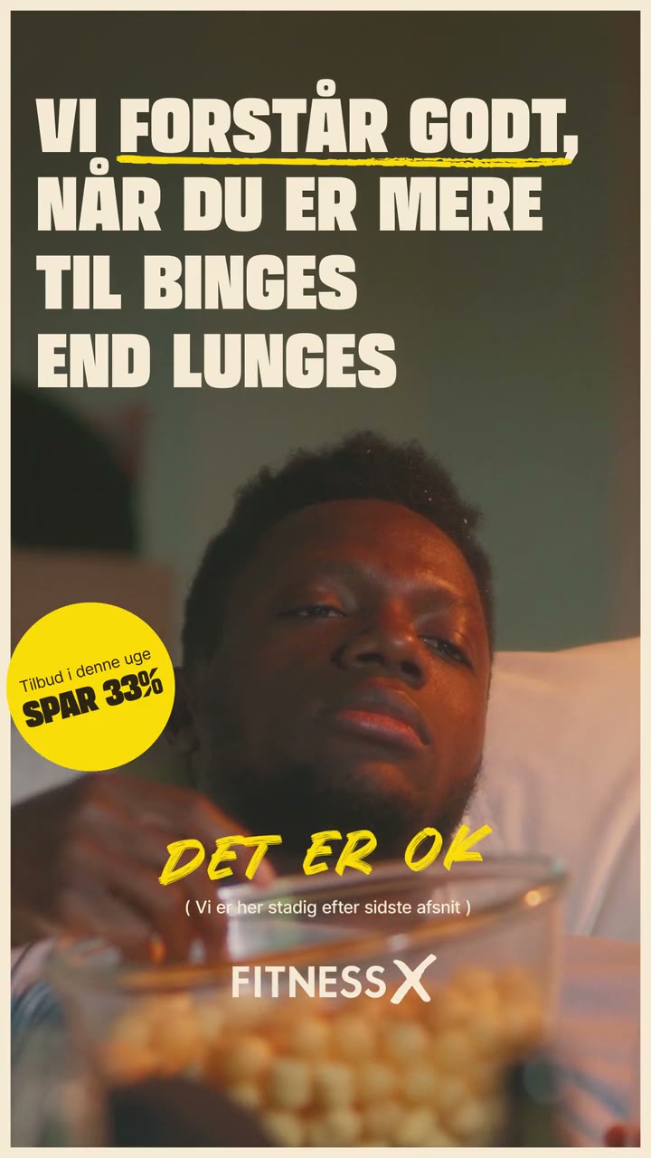 Vertical FitnessX ad: a man eats popcorn on the sofa under bold white type, "Vi forstår godt, når du er mere til binges end lunges", then "Det er OK" lands in yellow handwriting with a "Spar 33%" sticker and the FitnessX logo.